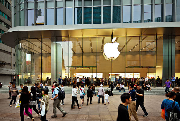 A bustling Apple Store with its iconic illuminated white Apple logo prominently displayed on the glass storefront. The modern retail location features floor-to-ceiling glass walls and is situated at street level of a contemporary urban building with reflective windows above. Large crowds of people gather outside and can be seen inside the brightly lit store. The scene shows typical busy foot traffic with shoppers of various ages walking by, some carrying bags, creating a vibrant street-level retail atmosphere. The architectural design emphasizes transparency and openness with extensive use of glass and clean geometric lines.