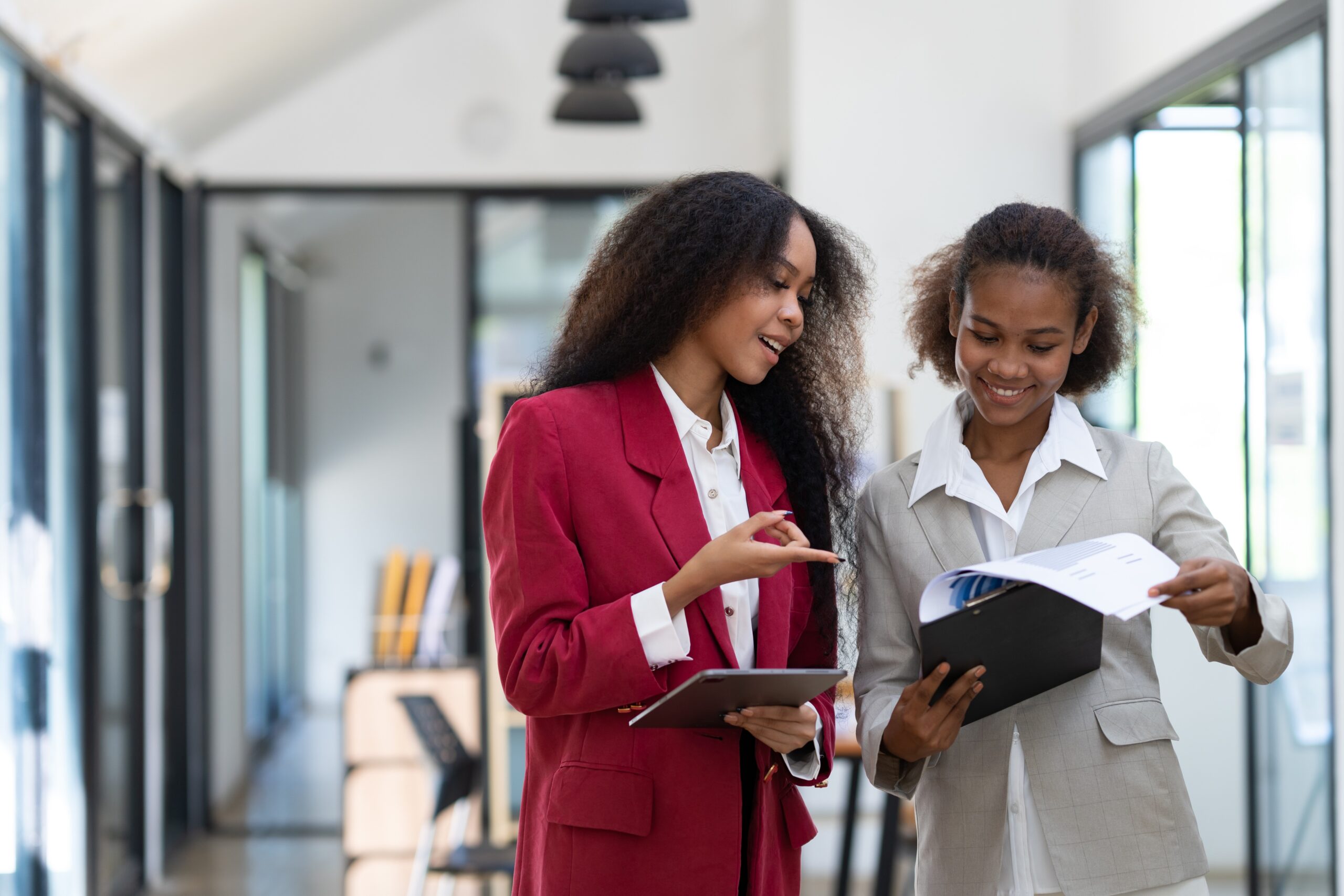 Two African American women in professional business attire walk together down a modern office hallway, reviewing documents and having a conversation. The woman on the left wears a red blazer over a white blouse and holds a tablet, while pointing at papers held by her colleague, who wears a beige/tan blazer over a white shirt. Both women are smiling as they collaborate. The hallway features floor-to-ceiling windows, white walls, and modern office furniture visible in the background