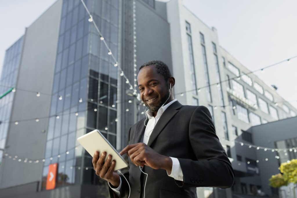 An African American businessman stands outdoors in an urban setting, smiling while holding a tablet and wearing earbuds. He is dressed professionally in a dark suit jacket over a white dress shirt. Behind him is a modern commercial building with glass facades and contemporary architecture. String lights are visible overhead, suggesting an outdoor plaza or pedestrian area. The setting appears to be a business district with modern office buildings, and the man's expression conveys confidence and professionalism as he appears to be engaged in mobile work or communication