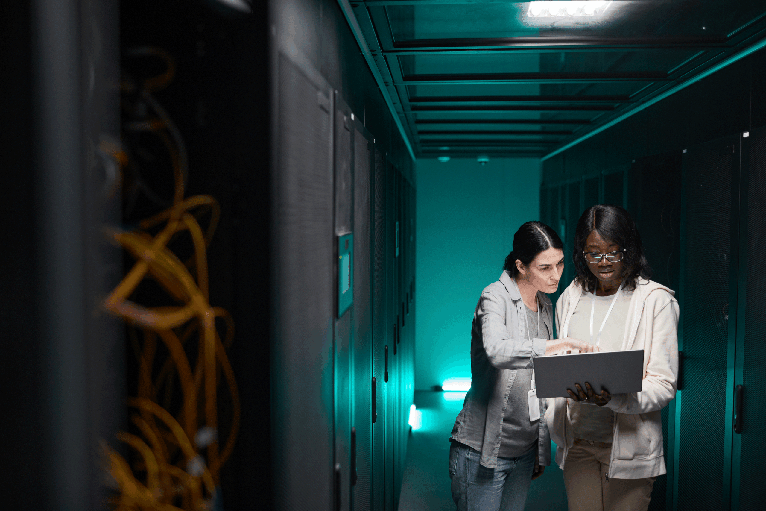 Two women, one African American and one Caucasian, stand together in a data center corridor examining a laptop computer. The Caucasian woman wears a gray hoodie and has dark hair pulled back, while the African American woman wears glasses and a white hoodie. They are surrounded by server racks in a hallway lit with teal/cyan colored lighting. Yellow cables are visible on equipment to the left, and the industrial setting suggests they are IT professionals or engineers working in a server facility.