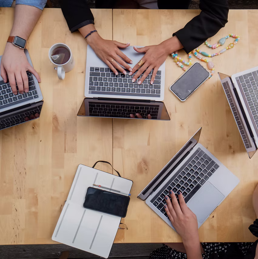 Top-down view of a collaborative workspace with diverse hands typing on laptops around a wooden table, surrounded by smartphones, notebooks, coffee, and accessories, symbolizing teamwork and digital productivity in a modern office setting