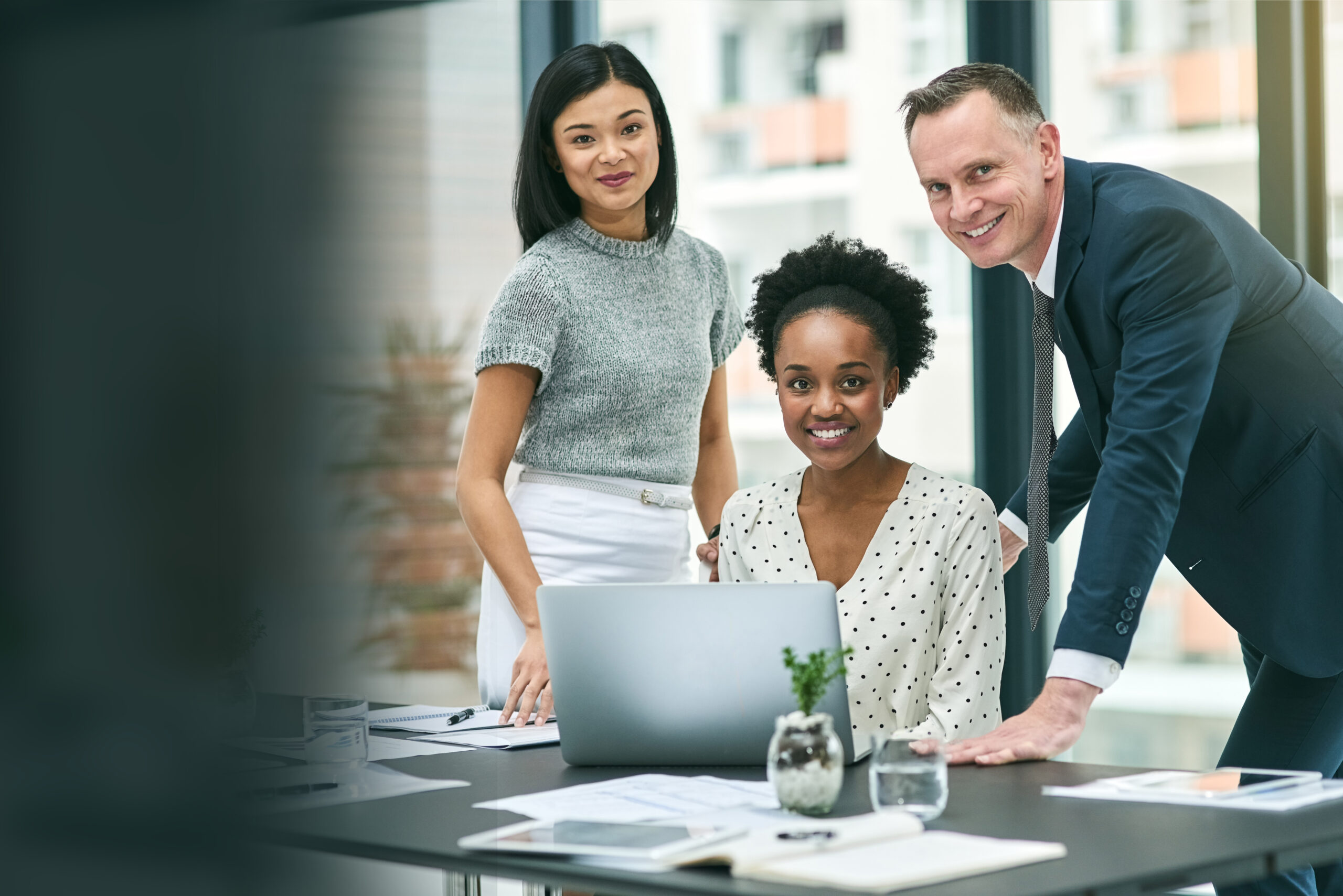 Three diverse business professionals—an Asian woman in a grey short-sleeve top and white skirt, a Black woman in a white polka dot blouse seated at a laptop, and a white man in a navy blue suit—collaborating and smiling during a meeting in a modern high-rise office with large glass windows, indoor plants, documents, and water glasses on the conference table